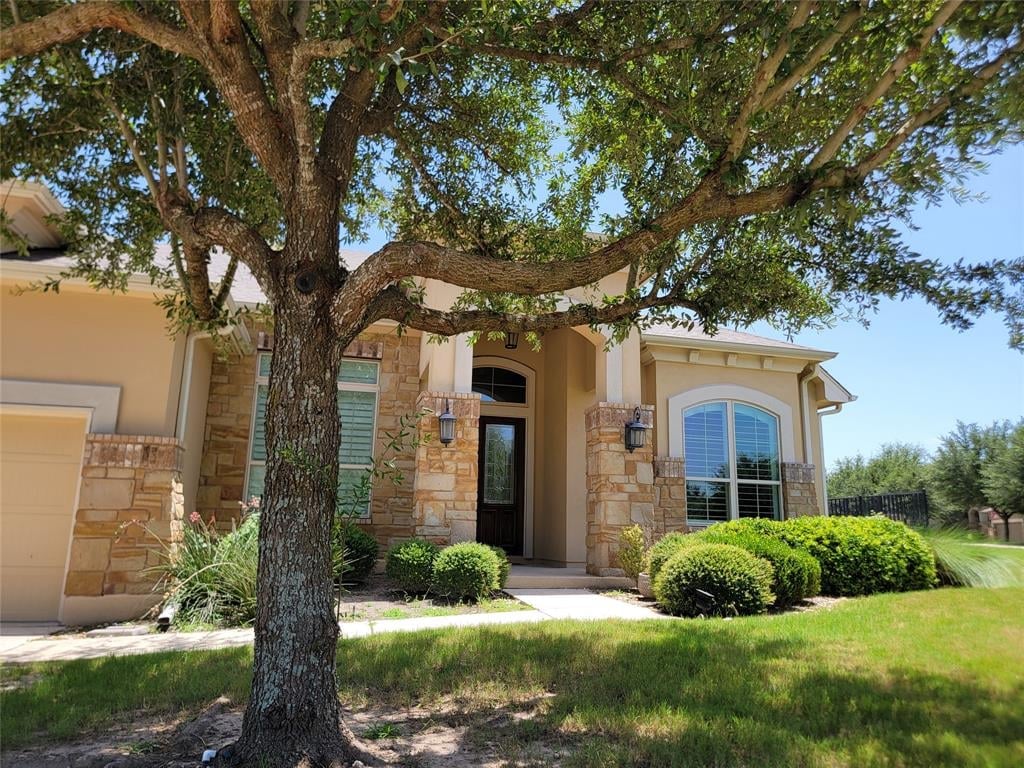 Modern stone house with tan exterior, arched windows, and large oak tree in front yard on sunny day