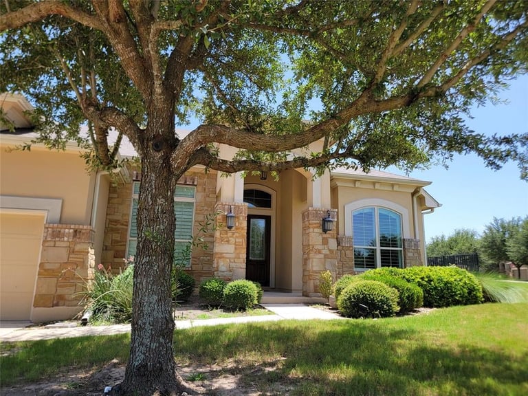Modern stone house with tan exterior, arched windows, and large oak tree in front yard on sunny day