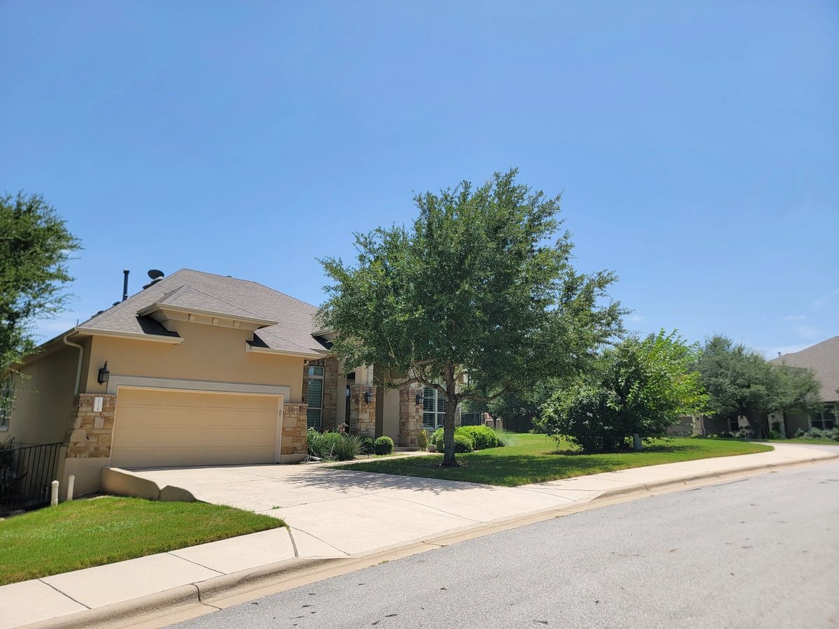 Suburban single-story home with tan brick exterior and garage on tree-lined residential street under clear blue sky
