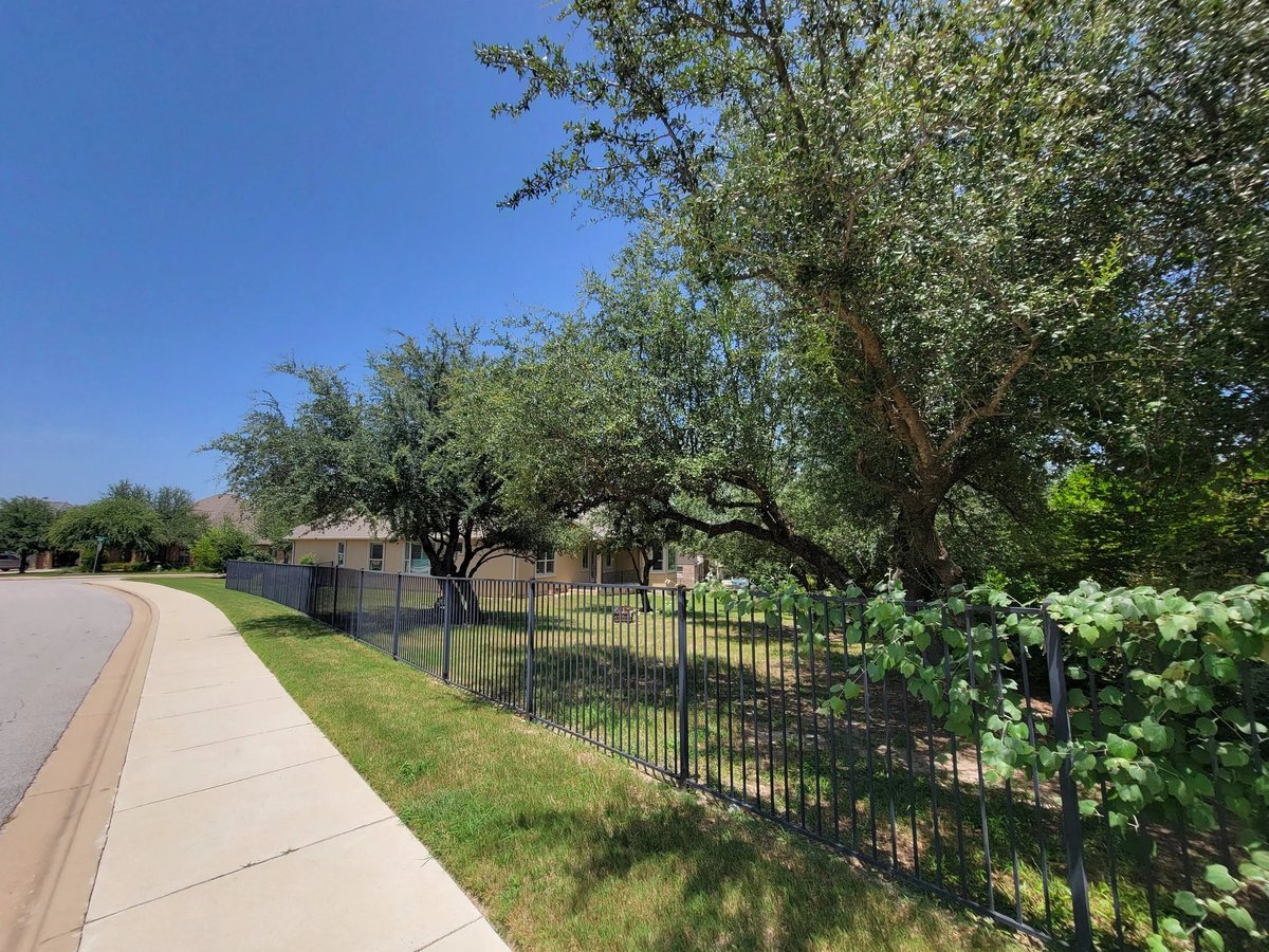Falconhead West neighborhood streetscape — wide sidewalks, black metal fencing, mature landscaping surrounding 16213 Maritime Alps Way in Austin TX. Lake Travis ISD schools nearby, minutes from Highway 71. Real Estates Of Texas, Gayle Sparkman listing. MLS 8675253