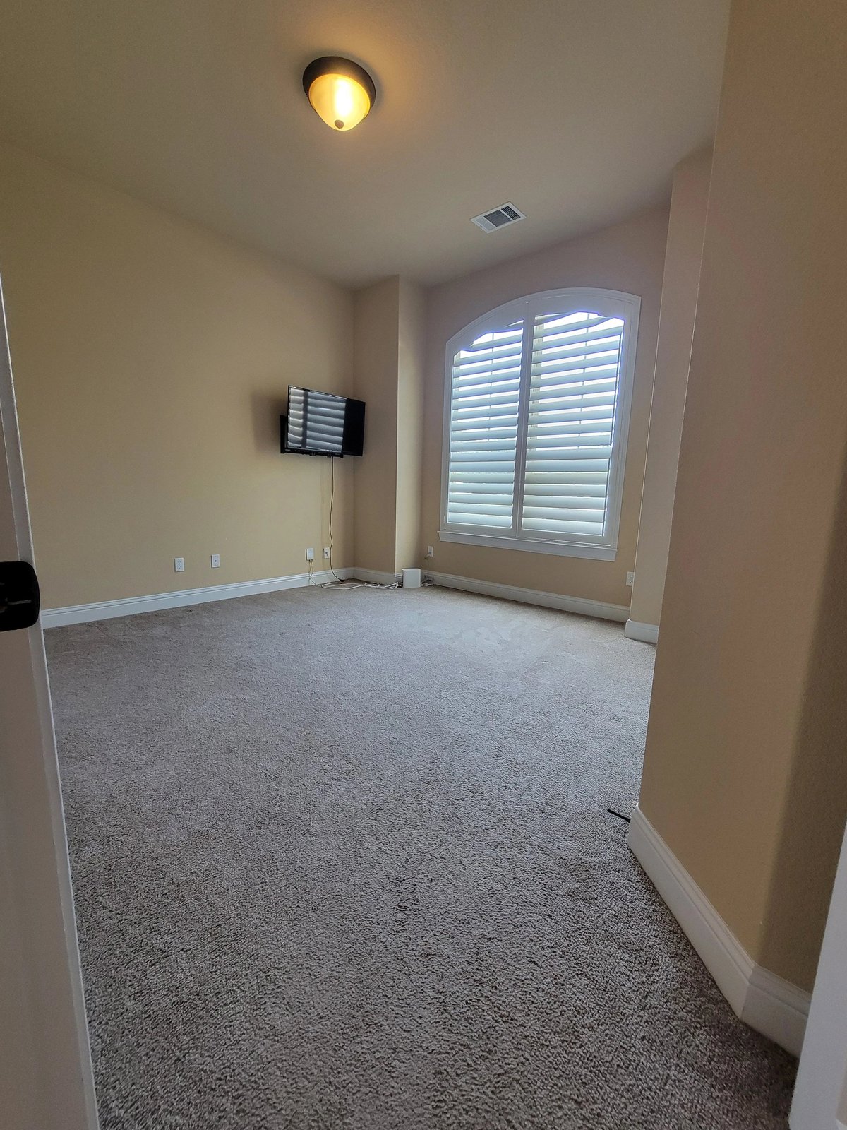 Empty room with beige walls, gray carpet, ceiling light fixture, white shuttered window, electrical outlet, and wall-mounted heater