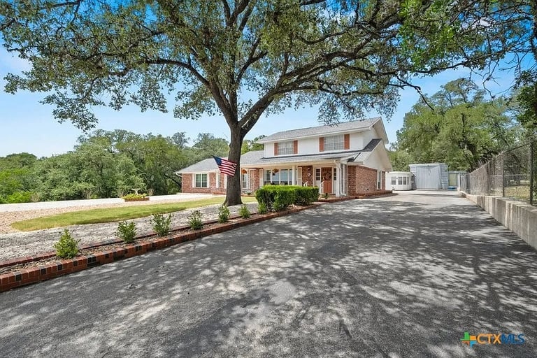 Brick two-story home with covered porch set on a tree-lined driveway with white fence and open grounds