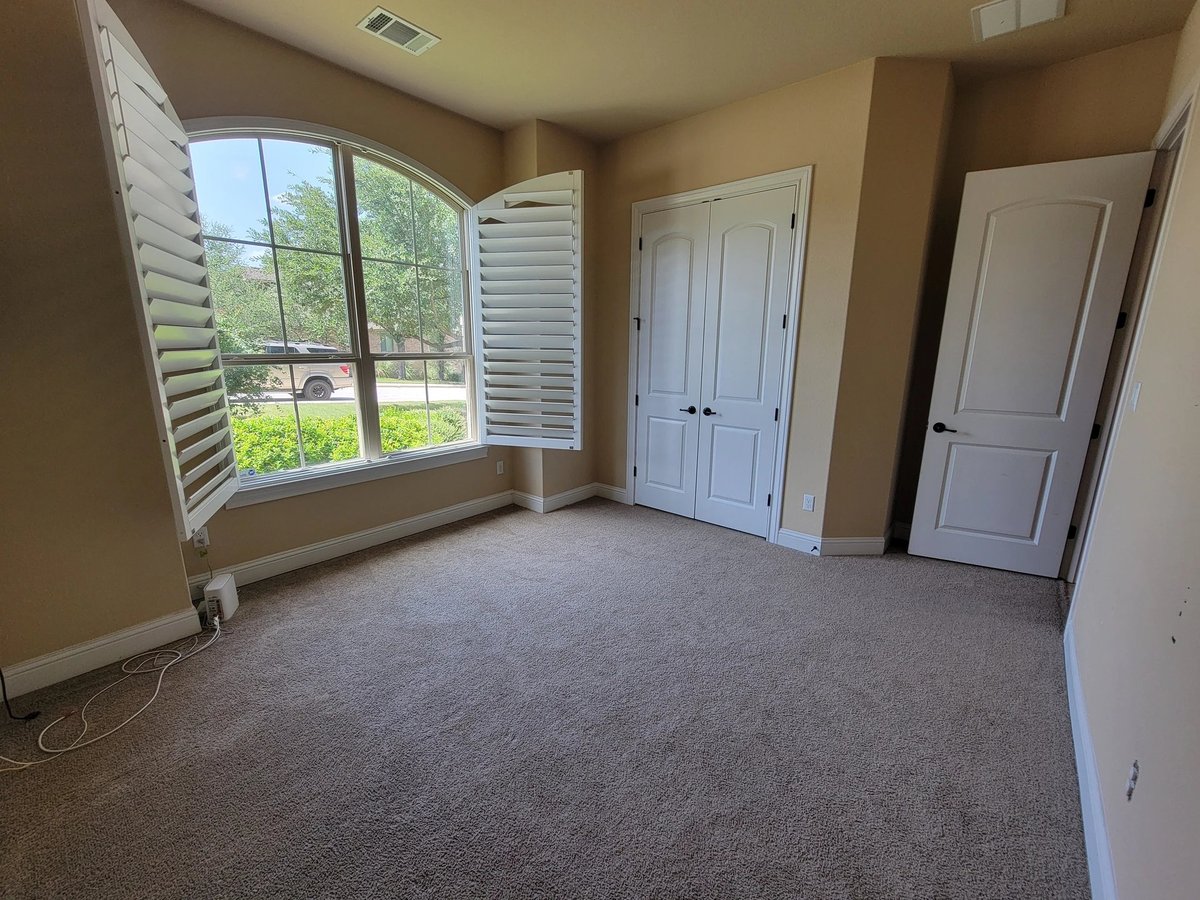 Empty bedroom with tan walls, gray carpet, arched window with shutters, and white closet doors