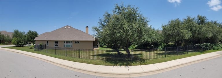 Single-story tan house with dark metal fence, mature trees in yard, and paved driveway on corner lot