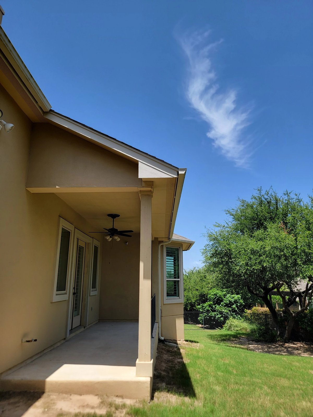 Tan house exterior with covered porch, glass doors, and adjacent green lawn with landscaping under blue sky