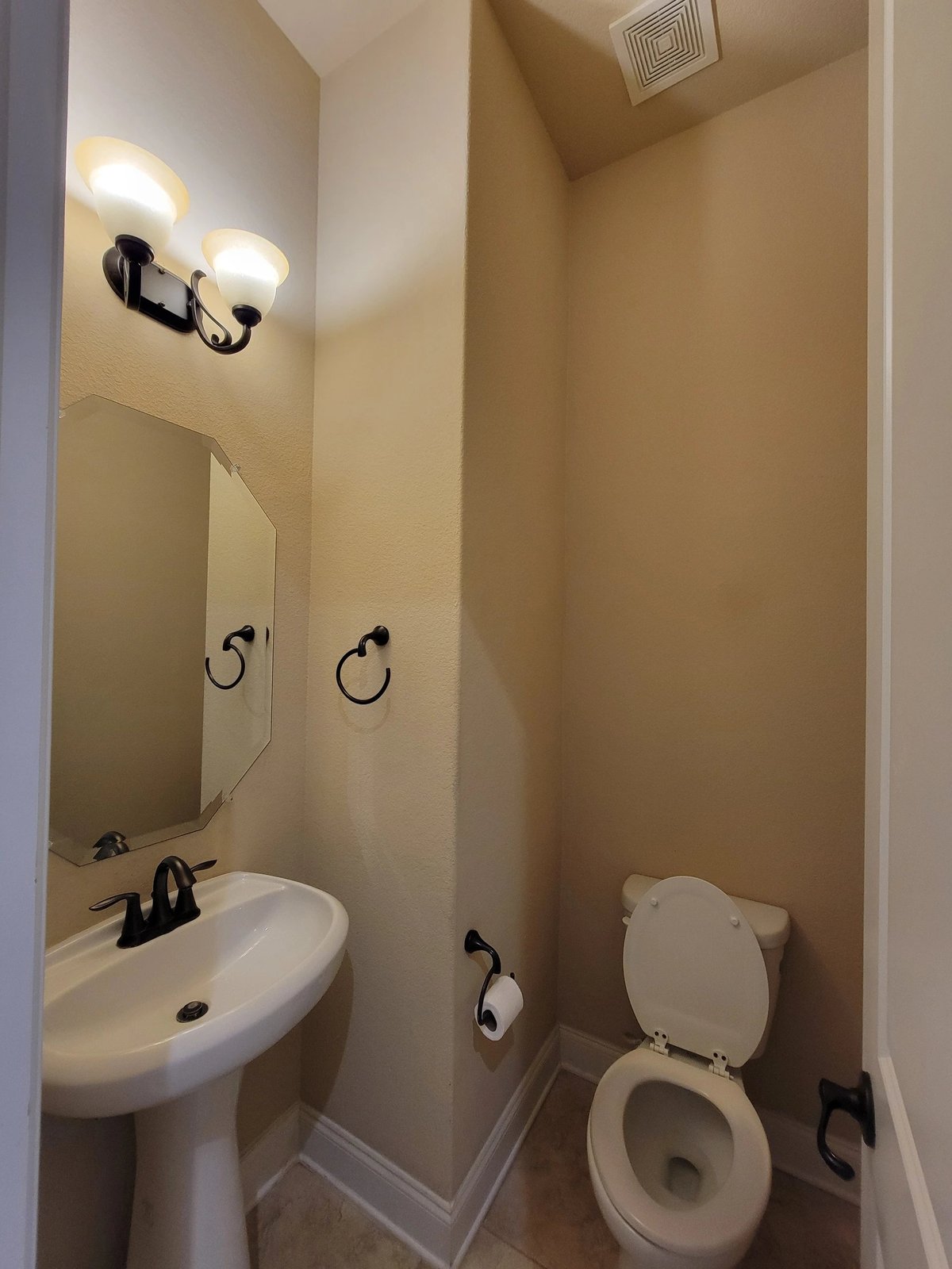 Small bathroom with beige walls, white pedestal sink, toilet, mirror with lighting, and towel rings