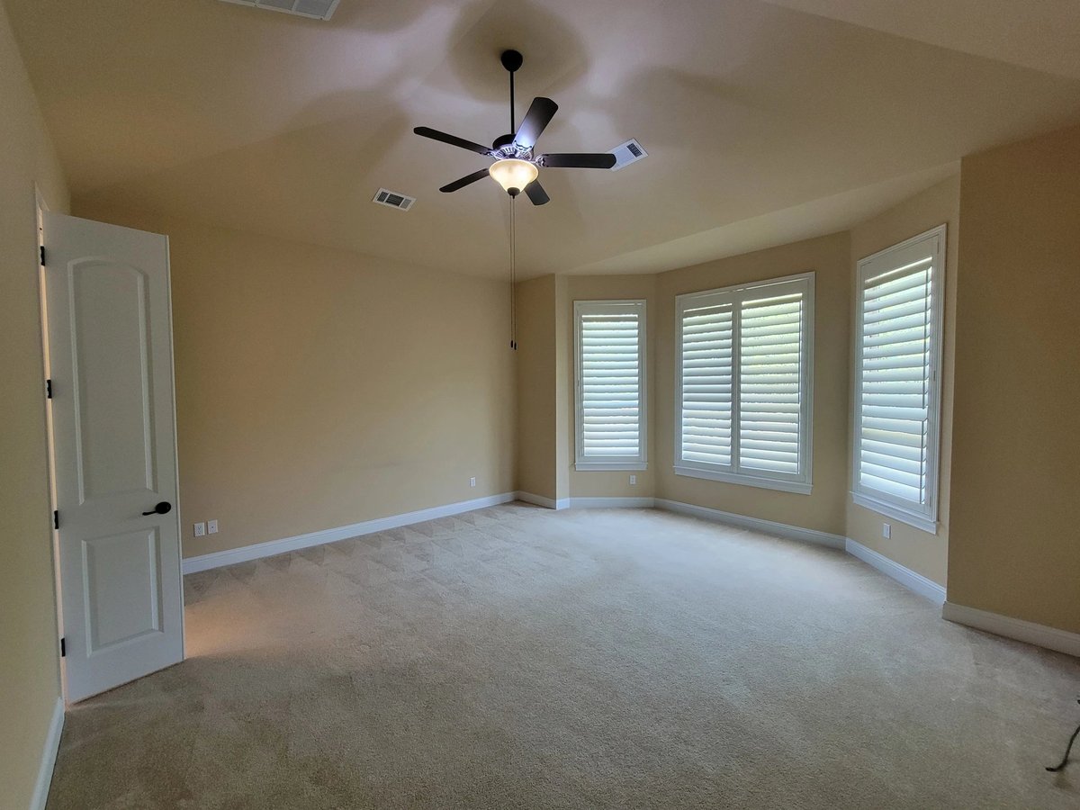 Empty bedroom with beige carpet, cream walls, ceiling fan, white shuttered windows, and an open door on the left wall