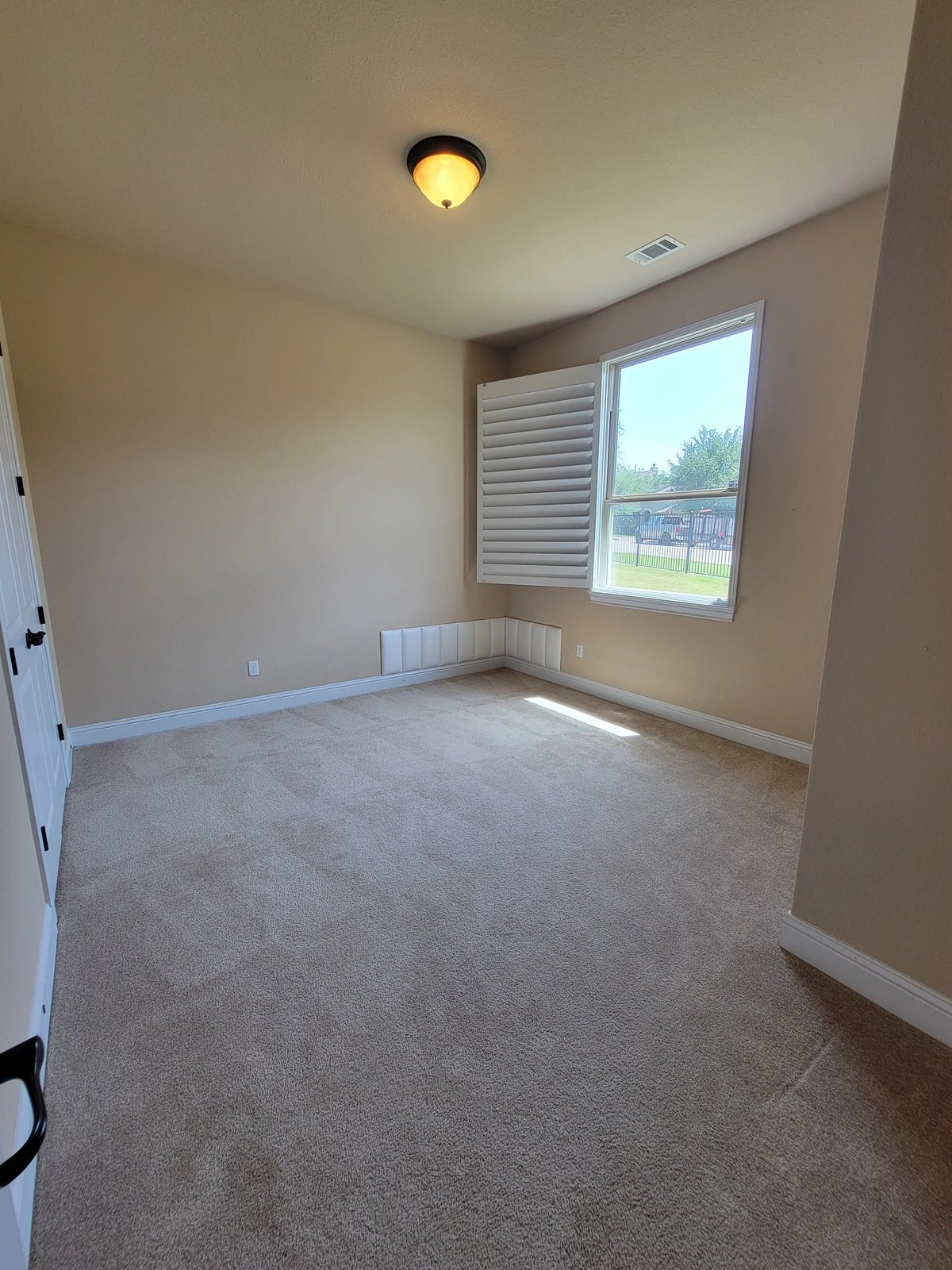 Empty bedroom with beige walls, carpet flooring, ceiling light, window with blinds, and door on the left