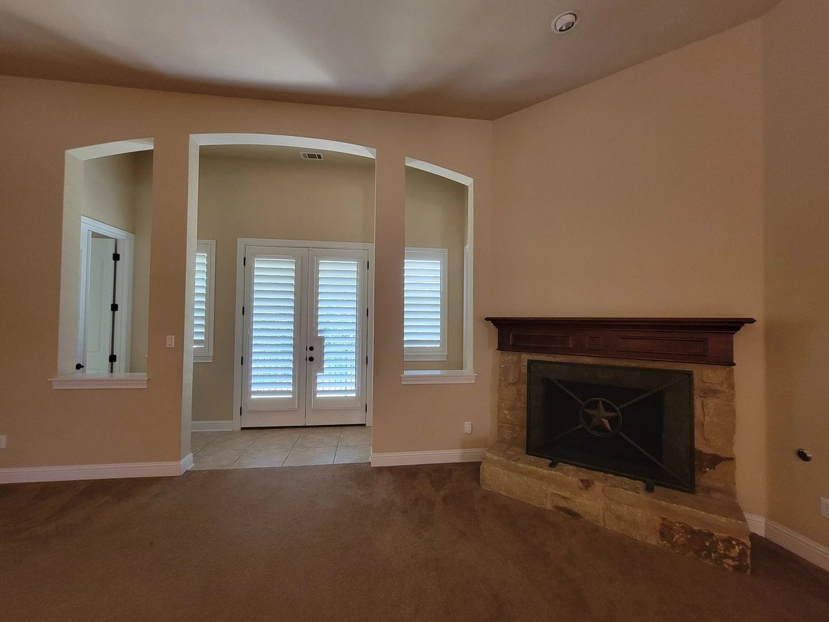 Empty room with cream walls, fireplace on right, and glass doors with shutters leading to patio