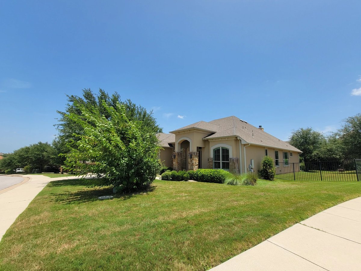 Single-story tan brick house with green lawn, large tree, and driveway under clear blue sky