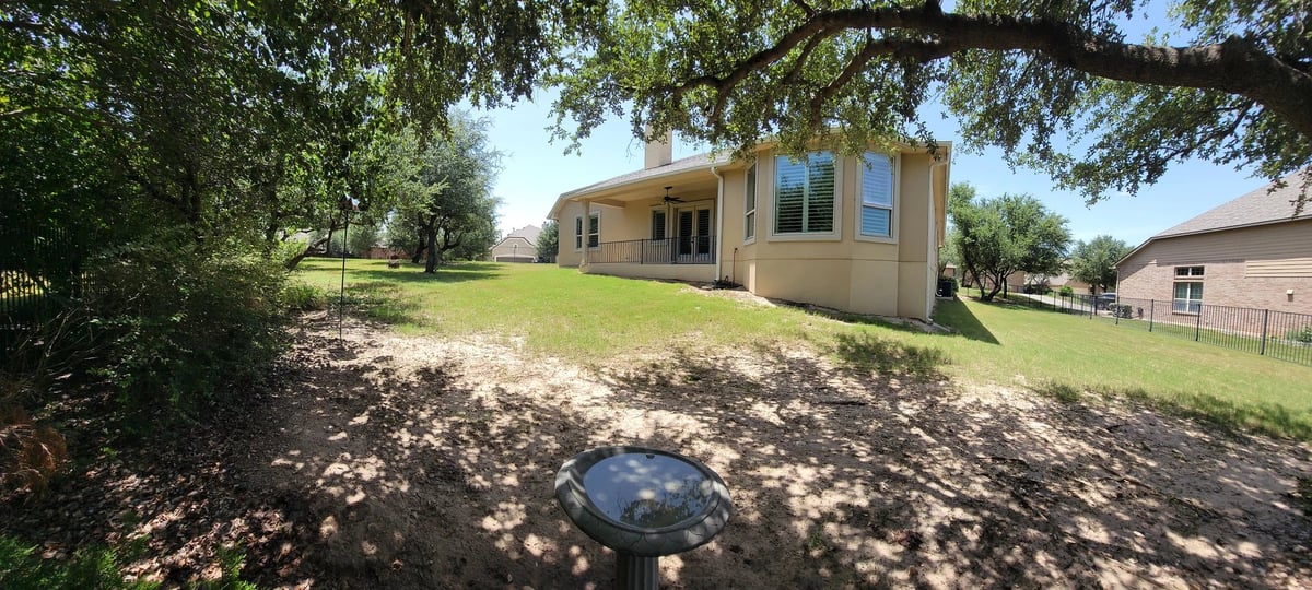 Tan colored house with blue shutters sitting on grassy lot under large oak trees on sunny day