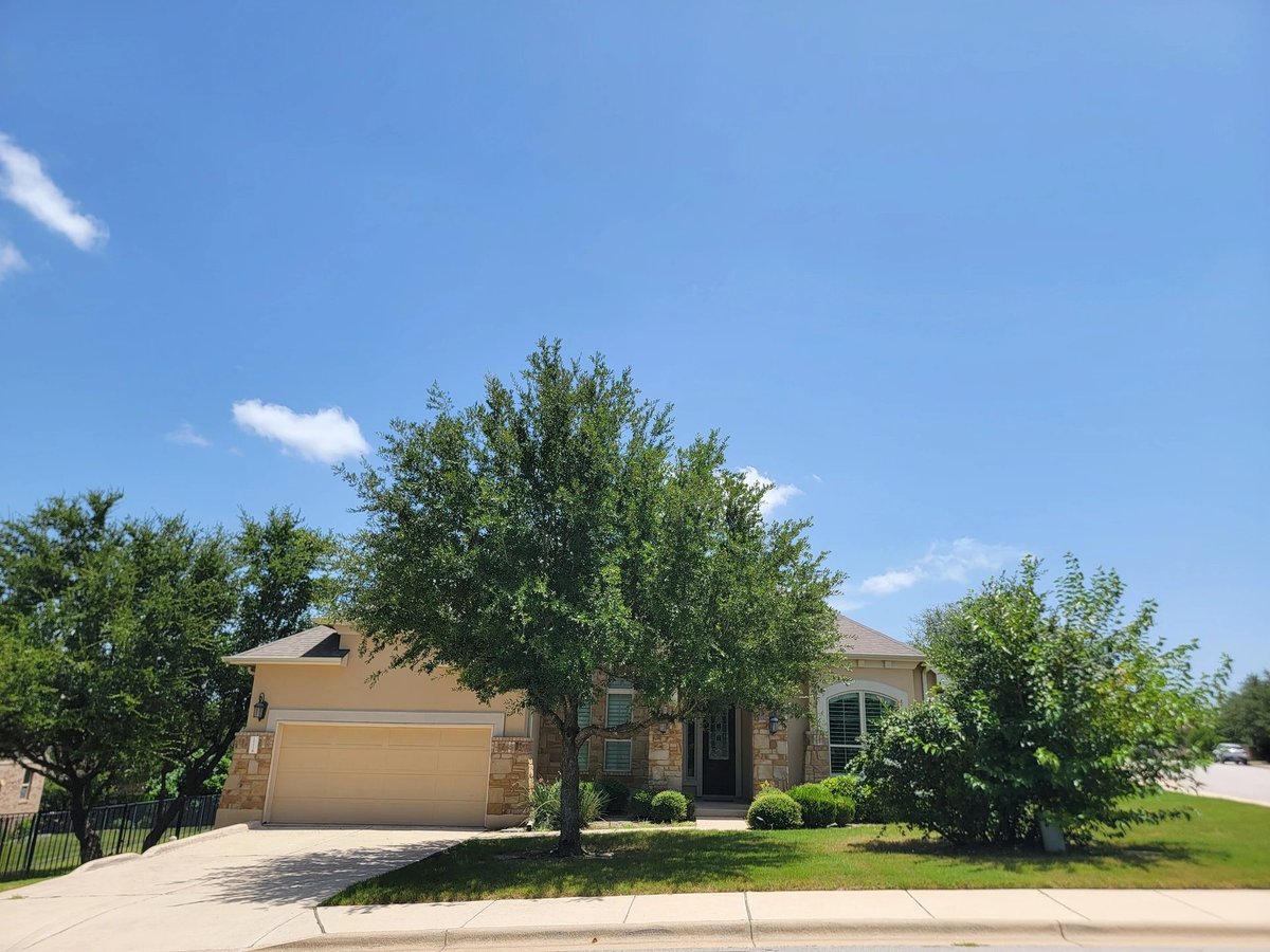 Front exterior of 16213 Maritime Alps Way — single-story beige brick home with garage, mature Live Oak trees, manicured lawn in Falconhead West, Austin TX 78738. 4BR 4BA 2,980 sqft corner lot. Listed by Gayle Sparkman, Real Estates Of Texas. MLS 8675253