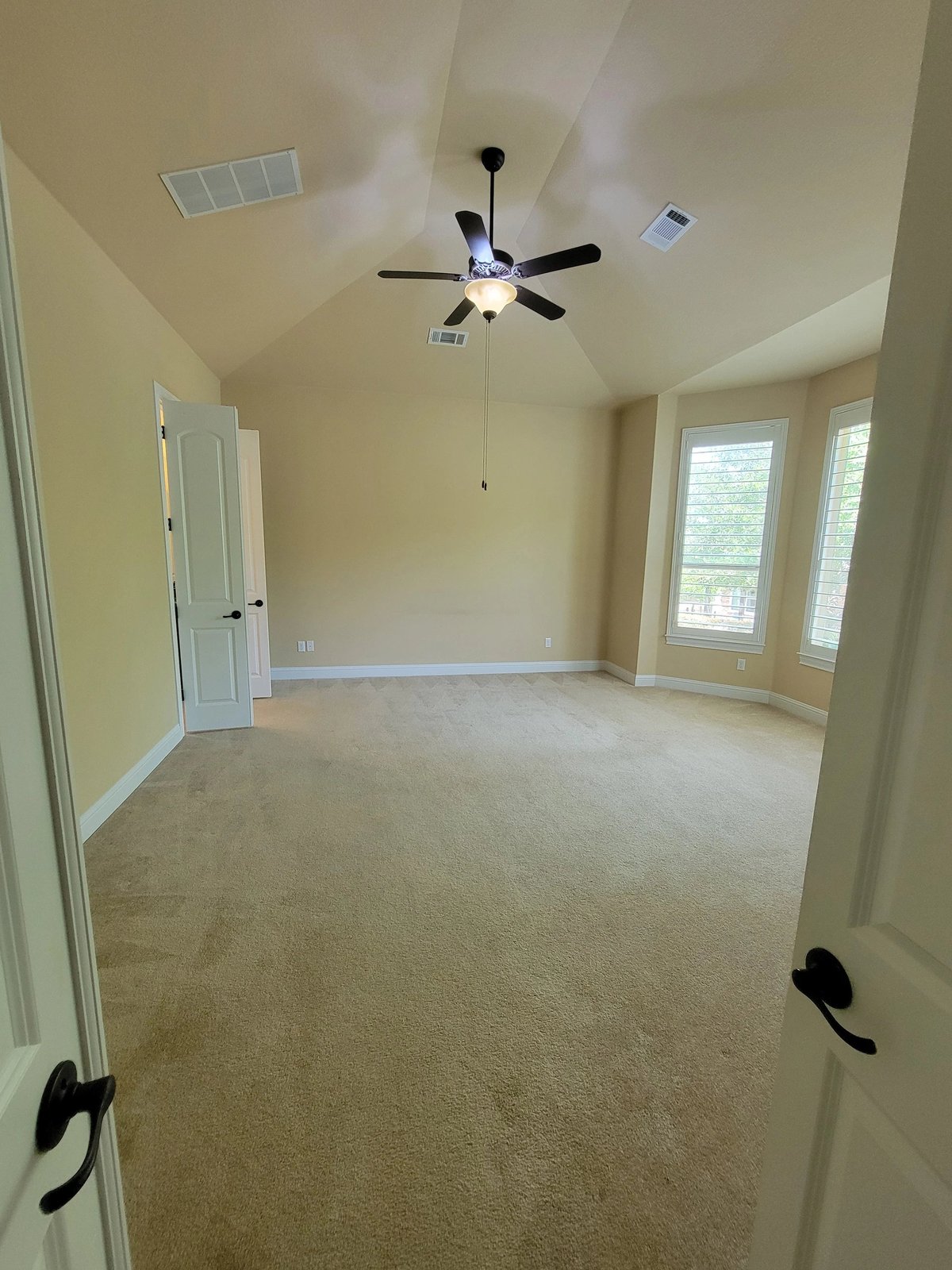 Empty room with beige carpet, ceiling fan, white walls, doors, and large windows with natural light