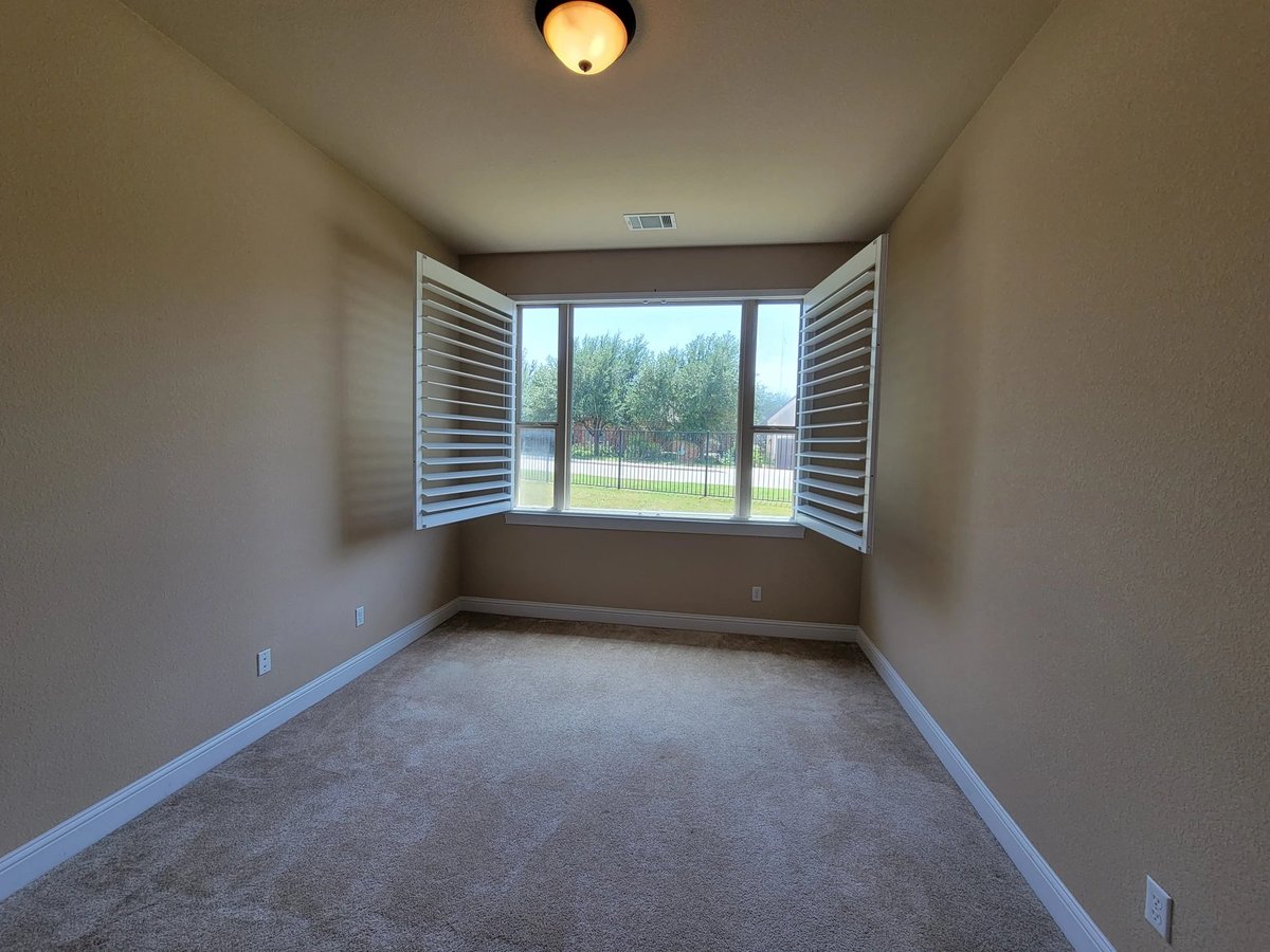 Empty room with gray carpet, beige walls, ceiling light, and large windows with open shutters overlooking an outdoor field