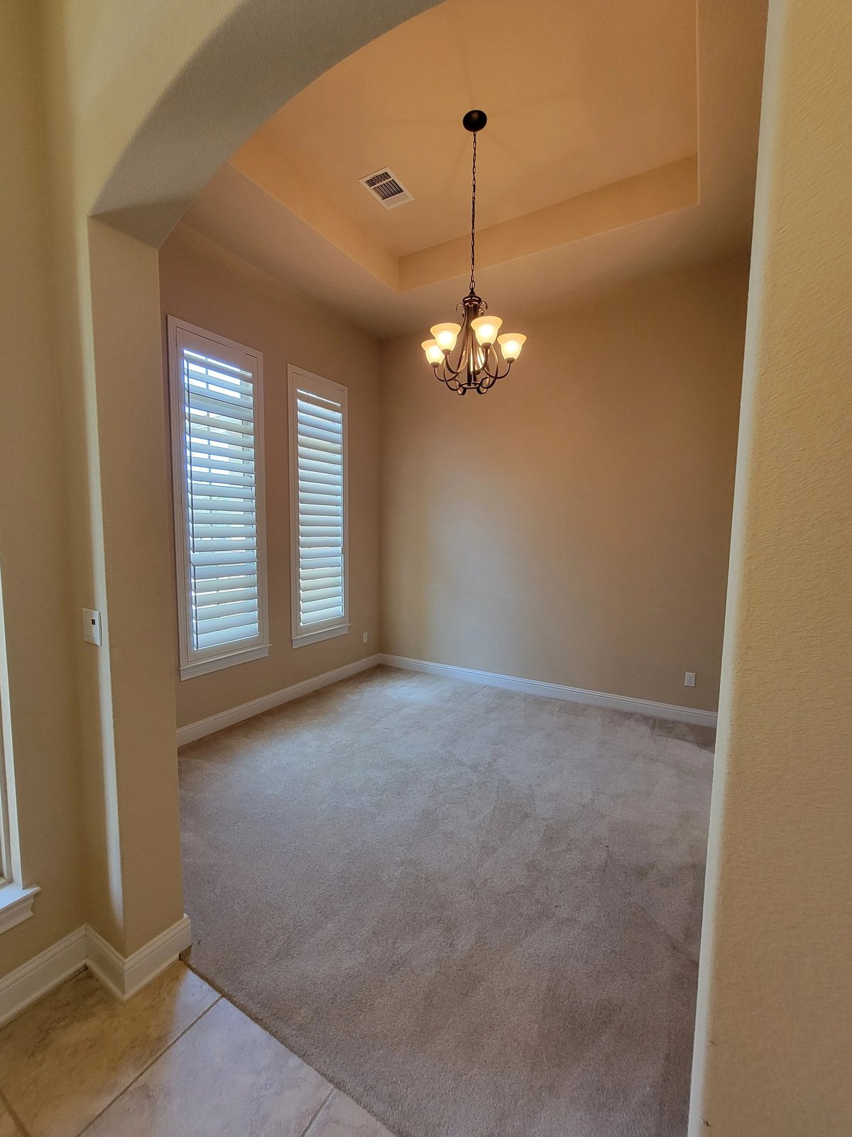 Empty room with beige walls, carpeted floor, pendant light fixture, and shuttered glass doors with natural lighting