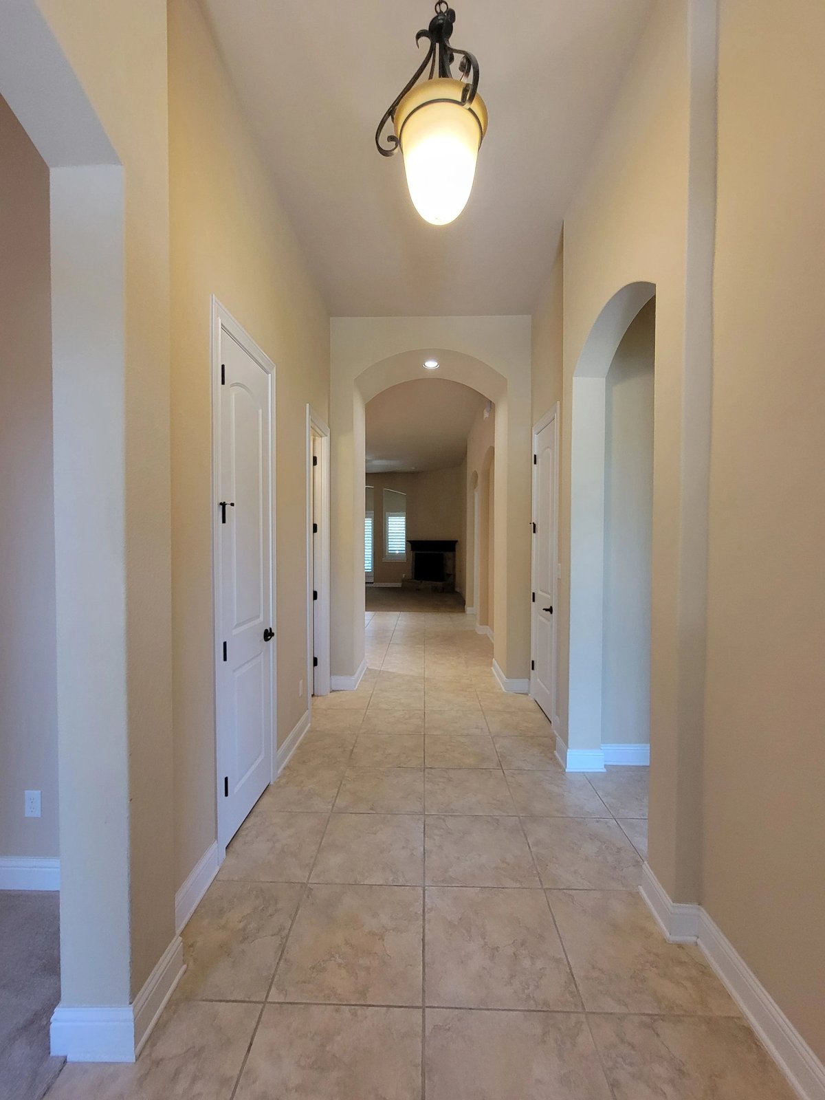 A long hallway with beige walls, tiled flooring, white doors on both sides, and a hanging pendant light fixture overhead