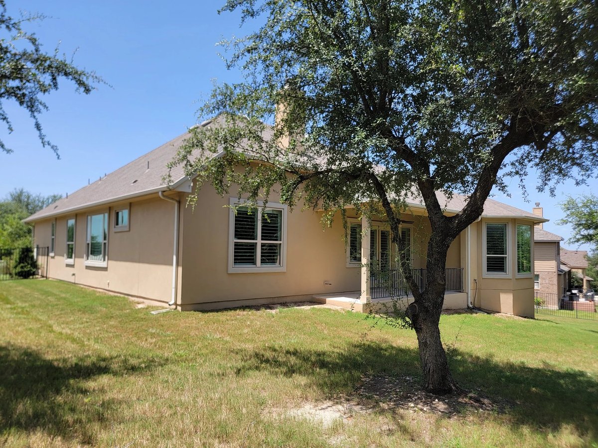 Single-story tan house with large tree in front yard on grassy lawn under clear blue sky