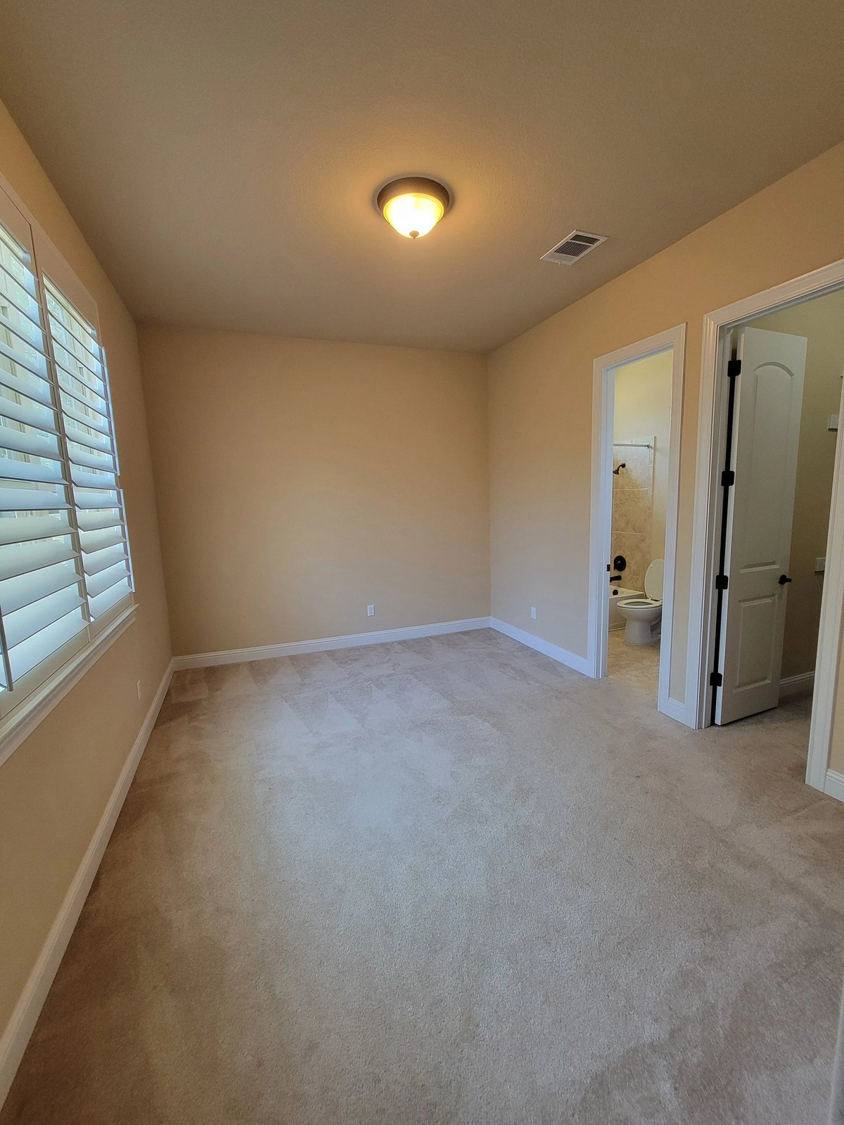 Empty room with beige walls, gray carpeting, ceiling light, window with shutters, and two open doorways leading to adjacent rooms