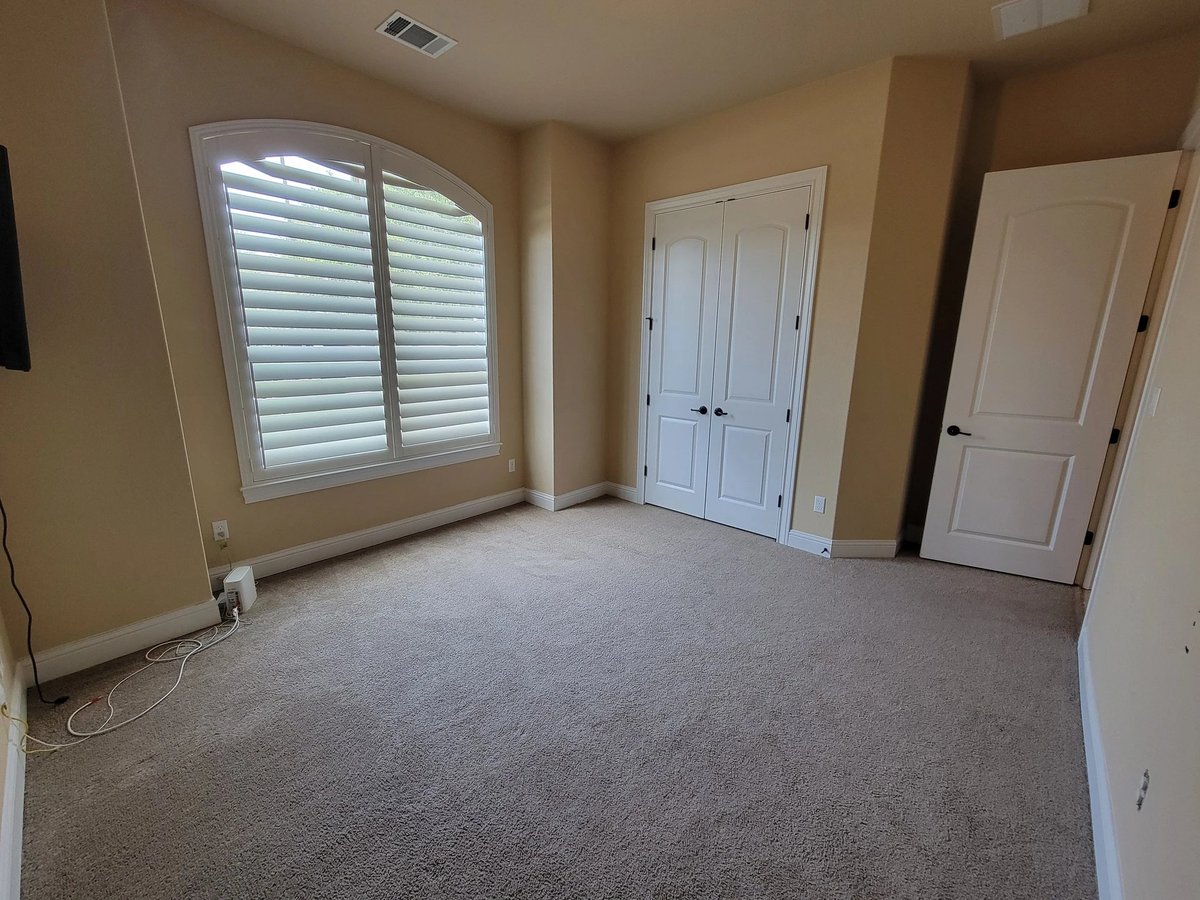 Empty beige bedroom with gray carpet, white shuttered window, and multiple white doors