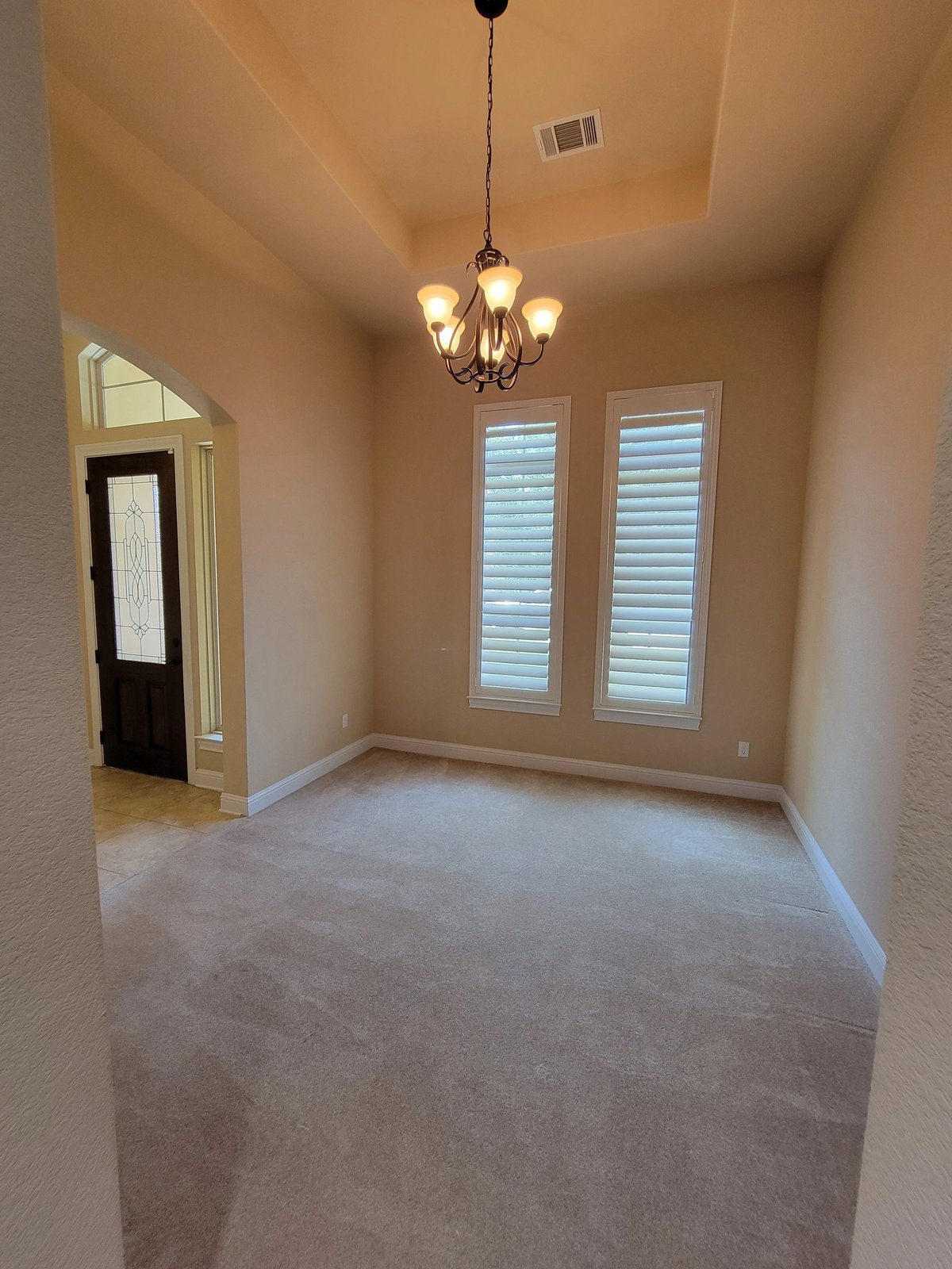 Empty room with beige walls, carpeted floor, hanging chandelier, and shuttered double doors and doorway